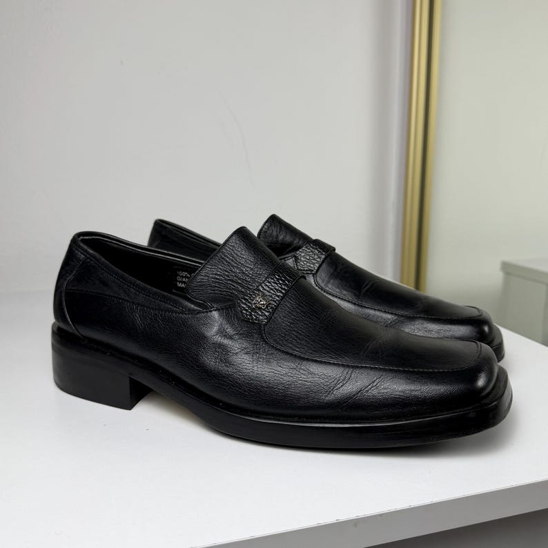 May include: A pair of black leather slip-on shoes with a square toe and a small block heel. The shoes have a decorative strap across the top with a silver-colored emblem. The shoes are displayed on a white surface.