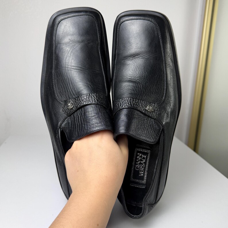 May include: Pair of black leather slip-on shoes with square toes. Each shoe features a textured strap with a small silver emblem. The insole of one shoe displays the "GIANNI VERSACE" brand name.