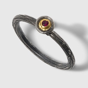 May include: A silver ring with a textured band and a gold-colored setting. The setting holds a small, round, red gemstone. The ring is displayed on a white background.