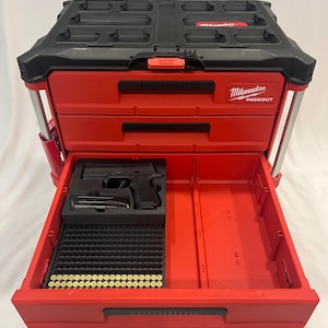 May include: Red and black Milwaukee Packout tool box with open drawer revealing a black handgun, magazine, and ammunition. The tool box has a black top and red drawers with the Milwaukee Packout logo.