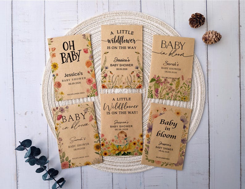 May include: A collection of baby shower seed packets with floral designs and text. The packets feature phrases like "Oh Baby" and "Baby in Bloom," along with names and dates. The packets are arranged on a woven placemat.