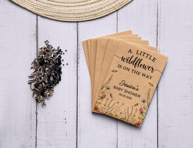 May include: A stack of kraft paper seed packets with the text "A little wildflower is on the way" and "Jessica's Baby Shower 08.09.2026". The packets are decorated with floral illustrations. A pile of seeds and a woven hat are also visible.
