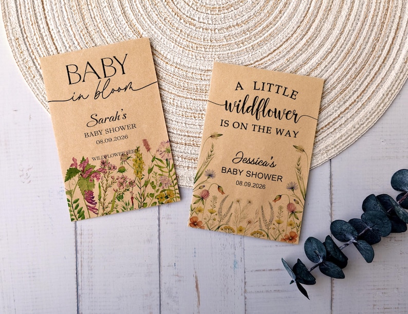 May include: Two kraft paper seed packets with floral designs and text. One reads "BABY in bloom" and "Sarah's BABY SHOWER 08.09.2026". The other says "A LITTLE wildflower IS ON THE WAY" and "Jessica's BABY SHOWER 08.09.2026".