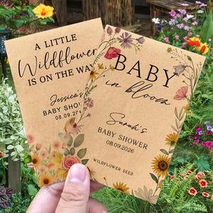 May include: Two kraft paper baby shower announcements with floral designs. One reads "A Little Wildflower Is On The Way," and the other says "Baby In Bloom." Both include baby shower details and wildflower seed information.