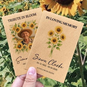 May include: Two seed packets with sunflower illustrations and text. One reads "TRIBUTE IN BLOOM" with a photo of a man in a hat. The other says "IN LOVING MEMORY" with a bouquet of sunflowers. The packets are held in front of a sunflower field.