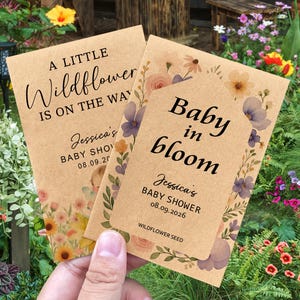 May include: Two kraft paper baby shower seed packets. One reads "A LITTLE Wildflower IS ON THE WAY" and the other says "Baby in bloom". Both packets are decorated with floral designs and include the baby shower details.