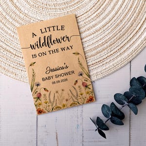 May include: A kraft paper baby shower invitation with the text "A little wildflower is on the way." The invitation includes the name "Jessica's" and the date "08.09.2026." Floral illustrations border the text. A sprig of eucalyptus is on the right.