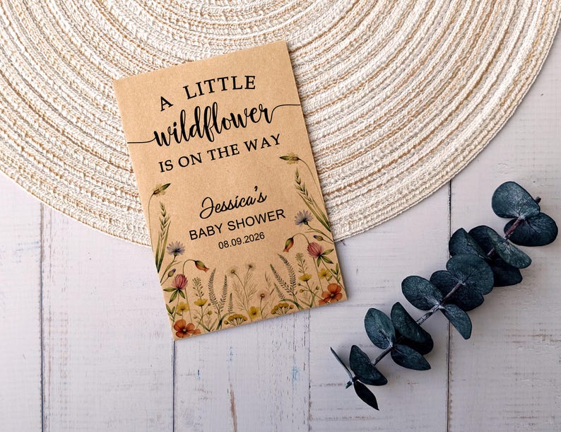 May include: A kraft paper baby shower invitation with the text "A little wildflower is on the way." The invitation includes the name "Jessica's" and the date "08.09.2026." Floral illustrations border the text. A sprig of eucalyptus is on the right.