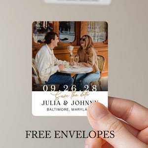 May include: A rectangular save the date card featuring a photograph of two people sitting at a cafe table. The card displays the date 09.26.28, the text Save the date, the names JULIA & JOHNNY, and the location BALTIMORE, MARYLAND in black and white.