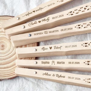 May include: A collection of personalized wooden fans, each engraved with names and dates. The fans are light-colored with intricate cut-out designs and silver rivets. The text includes names like "Sophia" and "Alice" with dates like "08.07.2026".