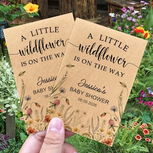May include: Two kraft paper baby shower announcements with floral illustrations. The text reads "A LITTLE wildflower IS ON THE WAY" and "Jessica's BABY SHOWER 08.09.2026". The announcements are held in front of a garden.