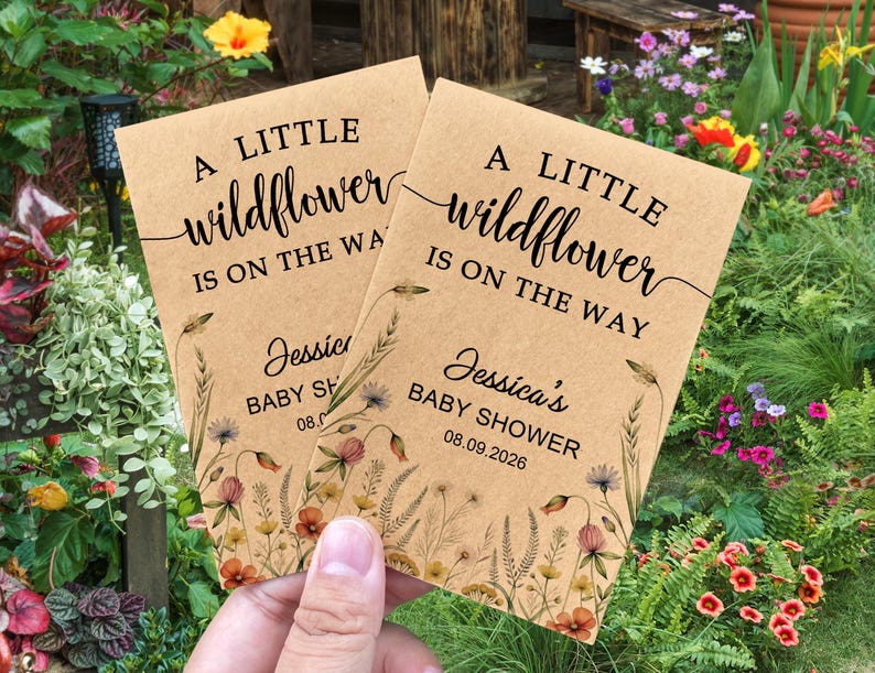 May include: Two kraft paper baby shower announcements with floral illustrations. The text reads "A LITTLE wildflower IS ON THE WAY" and "Jessica's BABY SHOWER 08.09.2026". The announcements are held in front of a garden.