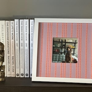 May include: A white framed picture with a pink and orange striped background, displaying a photo of a dog. The frame is on a shelf with books and a small statue. The books include "The Paris Review" series.