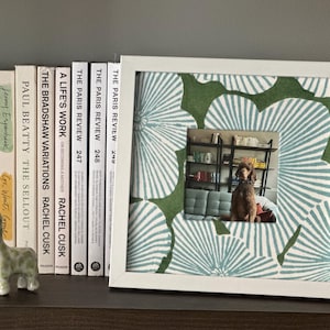 May include: A white framed picture with a green and white floral pattern, displaying a photo of a dog on a couch. The frame sits on a shelf with books and a giraffe figurine.