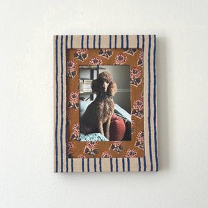 May include: A rectangular picture frame featuring a beige border with vertical navy blue stripes. The inner matting is a mustard yellow fabric decorated with a repeating pattern of small pink and green floral motifs, enclosing a photo of a brown poodle.