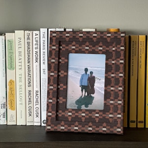 May include: A framed photograph with a brown and tan patterned border sits on a shelf with a collection of books. The photo shows two people on a beach. The books have white, yellow, and orange spines.