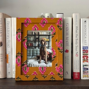 May include: A decorative photo frame with a mustard yellow background and a repeating floral pattern in pink and green. The frame holds a photo of a dog. Books are visible on either side of the frame.
