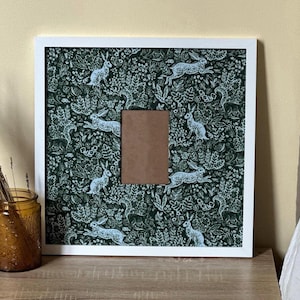 May include: Square white picture frame with a green and white rabbit pattern. The frame has a brown cardboard insert in the center. The background is a light beige color. A brown glass jar with dried plants is on the left.