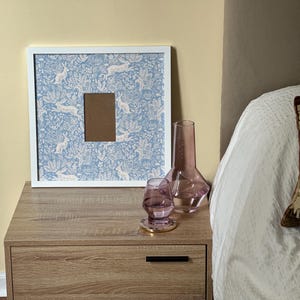 May include: A white framed picture with a blue and white rabbit pattern, a brown picture opening, and a decorative vase and glass on a wooden nightstand. A decorative pillow is on the bed.