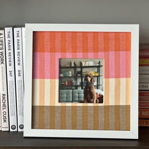 May include: A framed artwork featuring a central photograph of a dog in a room, surrounded by a striped pattern in shades of orange, pink, and tan. The frame is white, and books are visible on a shelf to the left.
