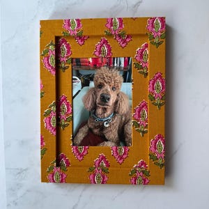 May include: A rectangular picture frame covered in mustard yellow fabric with a repeating pink and green floral pattern. The frame contains a photograph of a brown poodle with curly fur wearing a blue collar with a silver tag.