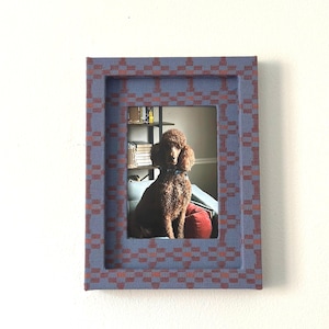 May include: A framed photograph of a brown poodle sitting on a blue couch with a red pillow. The frame is a rectangular shape with a blue and red checkered pattern.