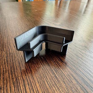 May include: A black, L-shaped 3D-printed object. The object has a textured surface and is designed with a series of parallel lines. The object is sitting on a wooden table.