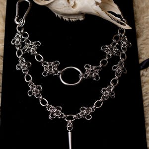 Peut inclure: Un collier en cotte de mailles argenté avec un grand anneau et un pendentif en forme de pointe. Le collier présente un motif floral et est présenté sur un fond noir, avec un crâne en arrière-plan.