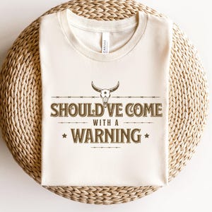 Könnte beinhalten: Ein weißes T-Shirt mit einer braunen Grafik eines Schädels mit Hörnern und dem Text "Should've Come With A Warning".
