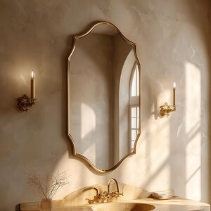 May include: A gold-framed mirror with an ornate, scalloped edge hangs above a marble vanity. Two gold sconces with lit candles flank the mirror. A small vase with dried stems sits on the vanity.