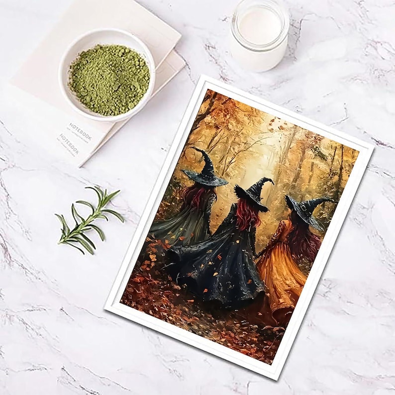 May include: A framed art print depicting three figures wearing pointed hats and long cloaks walking through an autumn forest with golden leaves. The print is displayed alongside a small white bowl of green powder and a sprig of rosemary.