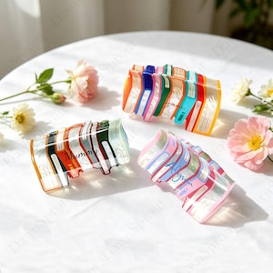 May include: Three colourful, translucent book-shaped acrylic bracelets. One bracelet has the word "Mummy" printed on it. Another has the name "Jane" and the third has the name "Alyssa". The bracelets have a striped design with various colours.