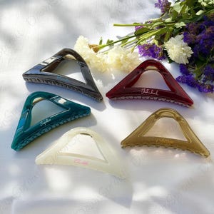 May include: Five triangular hair claw clips in various colors: dark gray, teal, burgundy, cream, and tan. Each clip has a gold-colored hinge and a name engraved on it: Sophie, Julilah, Adam, and Chloie. The clips are arranged on a white surface with flowers.