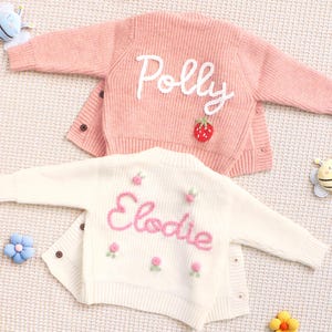 Könnte beinhalten: Zwei Baby-Strickjacken, eine rosa und eine cremefarbene, mit gestickten Namen und floralen Details. Die rosa Strickjacke trägt den Namen "Polly" in weißer Schrift und hat einen Erdbeerfleck. Die cremefarbene Strickjacke trägt den Namen "Elodie" in rosa Schrift und kleine rosa Blumen.