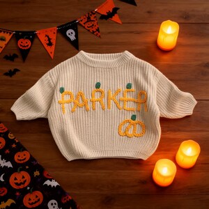 May include: A cream-coloured knitted sweater featuring the name "PARKER" in yellow, with green pumpkin details. The image includes Halloween decorations, suggesting a seasonal theme for children's clothing.