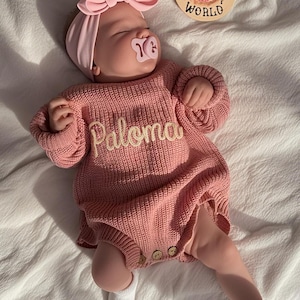 May include: A sleeping baby in a pink knitted romper with the name "Paloma" embroidered on the front. The baby wears a matching pink headband and has a pacifier. The romper has wooden buttons.