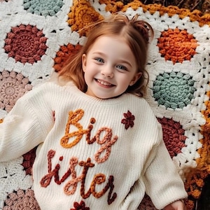 以下が含まれることがあります： オレンジと赤で「Big Sister」と刺繍されたオフホワイトのセーターを着た若い女の子が微笑んでいます。セーターには小さな花のアクセントがあります。彼女は、さまざまな花のパターンがあるカラフルなかぎ針編みの毛布の上に横たわっています。