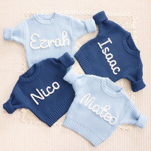 May include: Four long-sleeved knit sweaters in light blue and dark blue colors. Each sweater features a white embroidered name on the front, specifically the names Ezrah, Isaac, Nico, and Mateo, written in a cursive, textured yarn script.