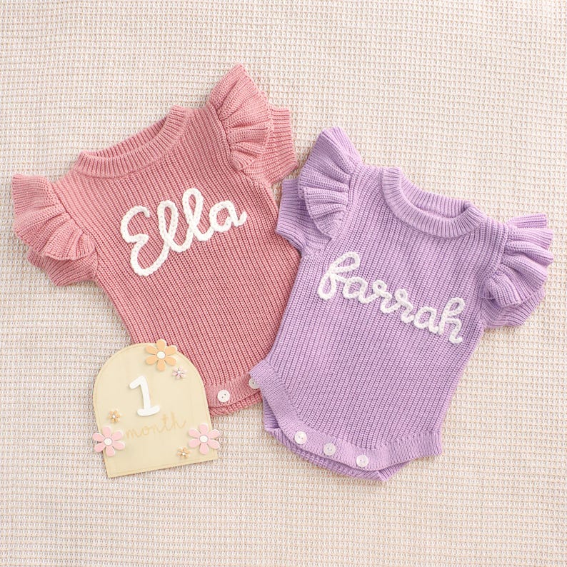 May include: Two baby bodysuits, one pink and one lavender, with ruffled sleeves and white embroidered names. A wooden milestone marker with the number "1" and floral accents is also present. The bodysuits are knit.