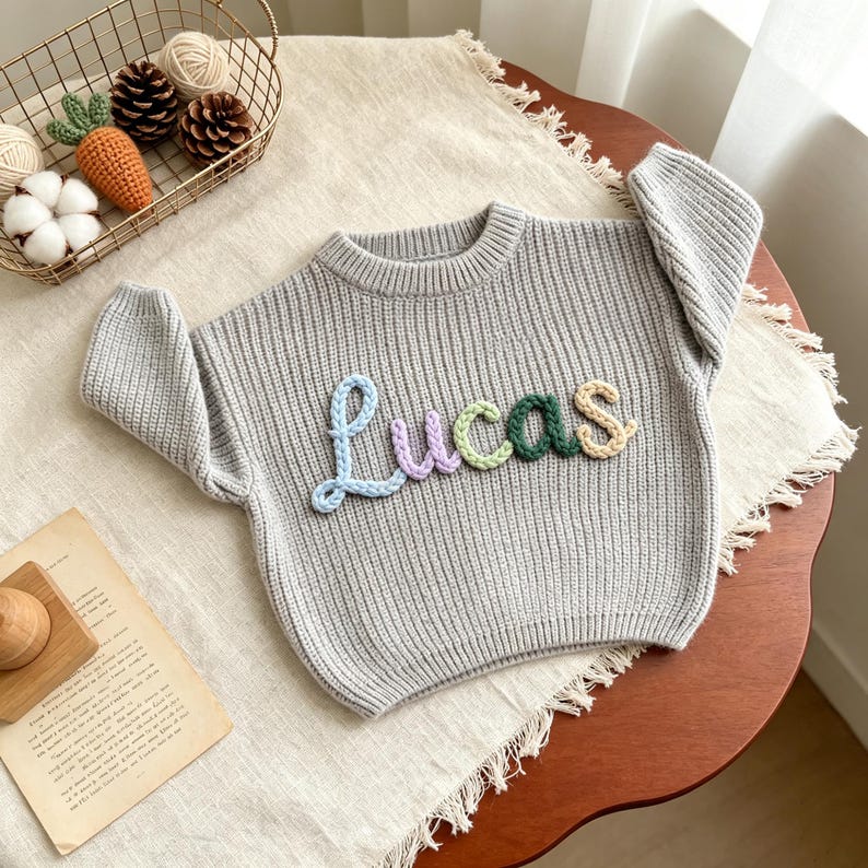 May include: A light gray long-sleeve knit sweater featuring the name Lucas embroidered in raised, multicolored yarn letters. The letters are light blue, lavender, light green, dark green, and beige, creating a textured, cursive appearance.
