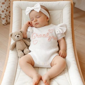 May include: A white knitted baby romper with ruffled sleeves, featuring the embroidered name Graelyn in pink cursive script and two small pink hearts. The romper is paired with a matching white headband with a large bow.