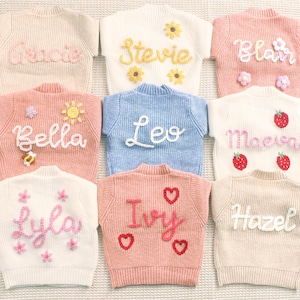 Peut inclure: Collection de neuf pulls pour enfants aux couleurs pastel. Chaque pull est orn&eacute; d'un pr&eacute;nom brod&eacute; en cursive, avec des &eacute;l&eacute;ments d&eacute;coratifs tels que des fleurs, des fraises et des c&oelig;urs. Les pulls sont dispos&eacute;s de mani&egrave;re ordonn&eacute;e, mettant en valeur les motifs personnalis&eacute;s.