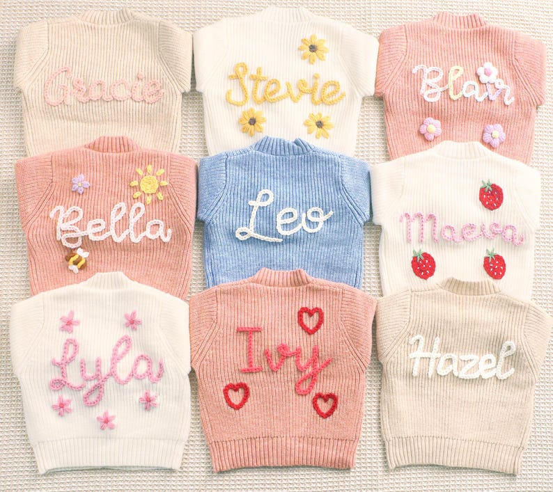 Peut inclure: Collection de neuf pulls pour enfants aux couleurs pastel. Chaque pull est orn&eacute; d'un pr&eacute;nom brod&eacute; en cursive, avec des &eacute;l&eacute;ments d&eacute;coratifs tels que des fleurs, des fraises et des c&oelig;urs. Les pulls sont dispos&eacute;s de mani&egrave;re ordonn&eacute;e, mettant en valeur les motifs personnalis&eacute;s.
