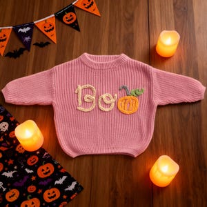 Op de afbeelding: Een roze gebreide trui met het woord "Boo" en een pompoen applicatie. De trui is versierd met een Halloween thema, met een banner van pompoenen en vleermuizen op de achtergrond. Kaarsen zijn rond de trui geplaatst.