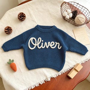 May include: A dark blue long-sleeve sweater featuring the name Oliver in white cursive yarn lettering across the chest. The sweater has a ribbed crew neckline, ribbed cuffs, and a ribbed hem, constructed with a chunky knit texture throughout.