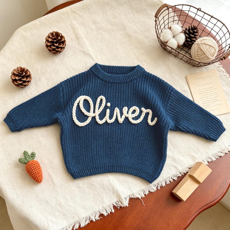 May include: A dark blue long-sleeve sweater featuring the name Oliver in white cursive yarn lettering across the chest. The sweater has a ribbed crew neckline, ribbed cuffs, and a ribbed hem, constructed with a chunky knit texture throughout.