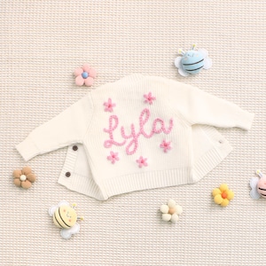Peut inclure: Cardigan de b&eacute;b&eacute; cr&egrave;me, brod&eacute; du pr&eacute;nom "Lyla" en rose, entour&eacute; de motifs de fleurs roses. Le cardigan a des boutons et est entour&eacute; d'ornements d&eacute;coratifs en forme de fleurs et d'abeilles.