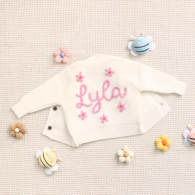 Peut inclure: Cardigan de b&eacute;b&eacute; cr&egrave;me, brod&eacute; du pr&eacute;nom "Lyla" en rose, entour&eacute; de motifs de fleurs roses. Le cardigan a des boutons et est entour&eacute; d'ornements d&eacute;coratifs en forme de fleurs et d'abeilles.