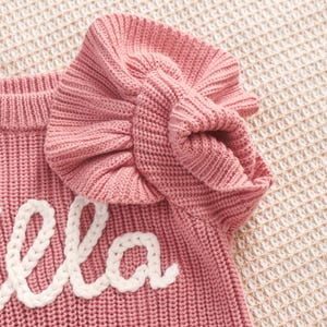 May include: A close-up of a pink knitted sweater with a ruffled sleeve. The word "ella" is embroidered in white cursive on the front of the sweater. The sweater is made of a soft, textured knit fabric.