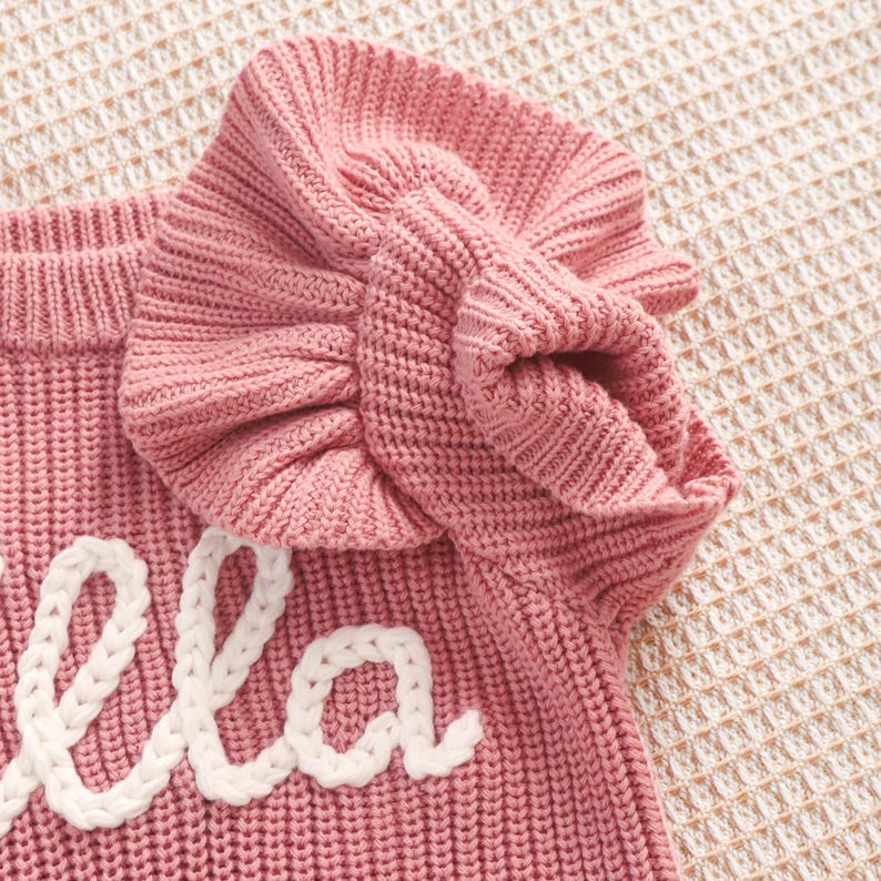 May include: A close-up of a pink knitted sweater with a ruffled sleeve. The word "ella" is embroidered in white cursive on the front of the sweater. The sweater is made of a soft, textured knit fabric.